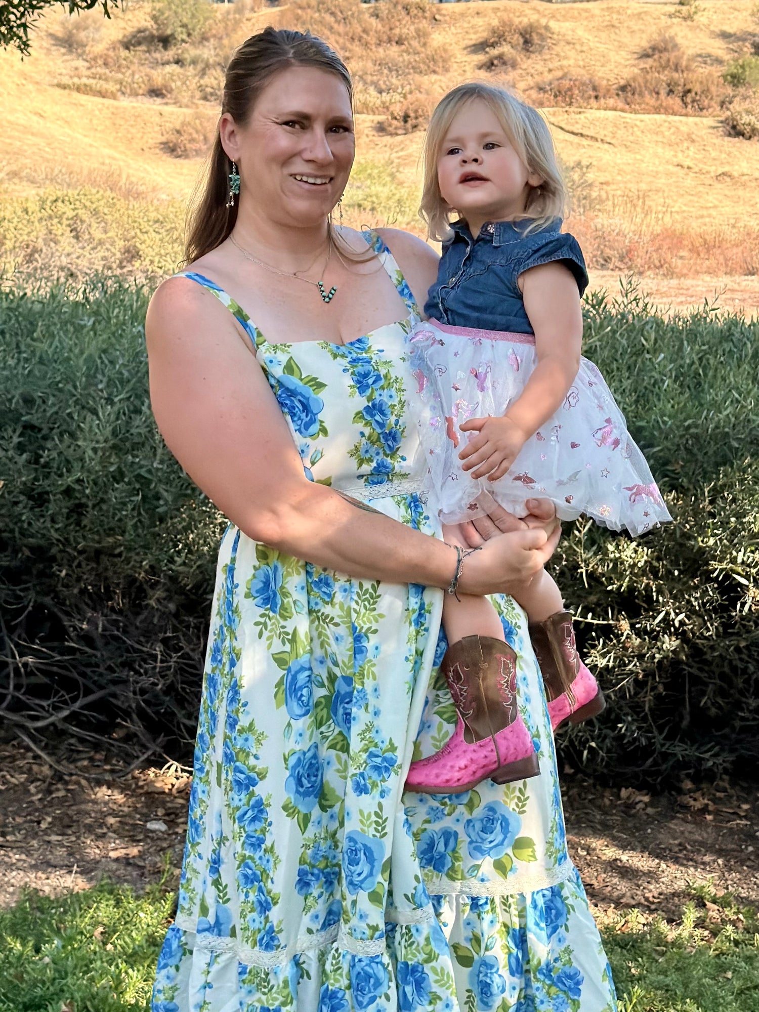 Stephanie in a blue and green floral sleeveless maxi dress holds Ry outdoors in front of a hedge and dry grassy hill.