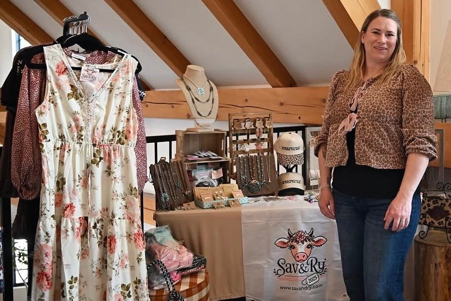 Stephanie Voss standing in a room with wooden beams and a table with a 'Sav & Ry' sign, surrounded by clothing and decor items.